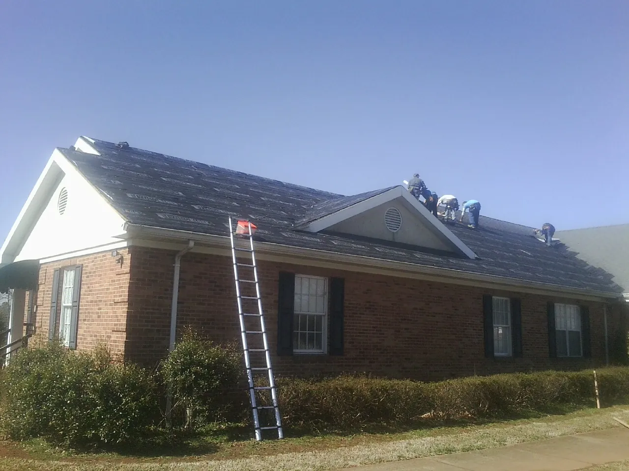 Crew tearing shingles off a brick ranch roof with a ladder set — Emergency Roof Repair work by our crew in New Glarus