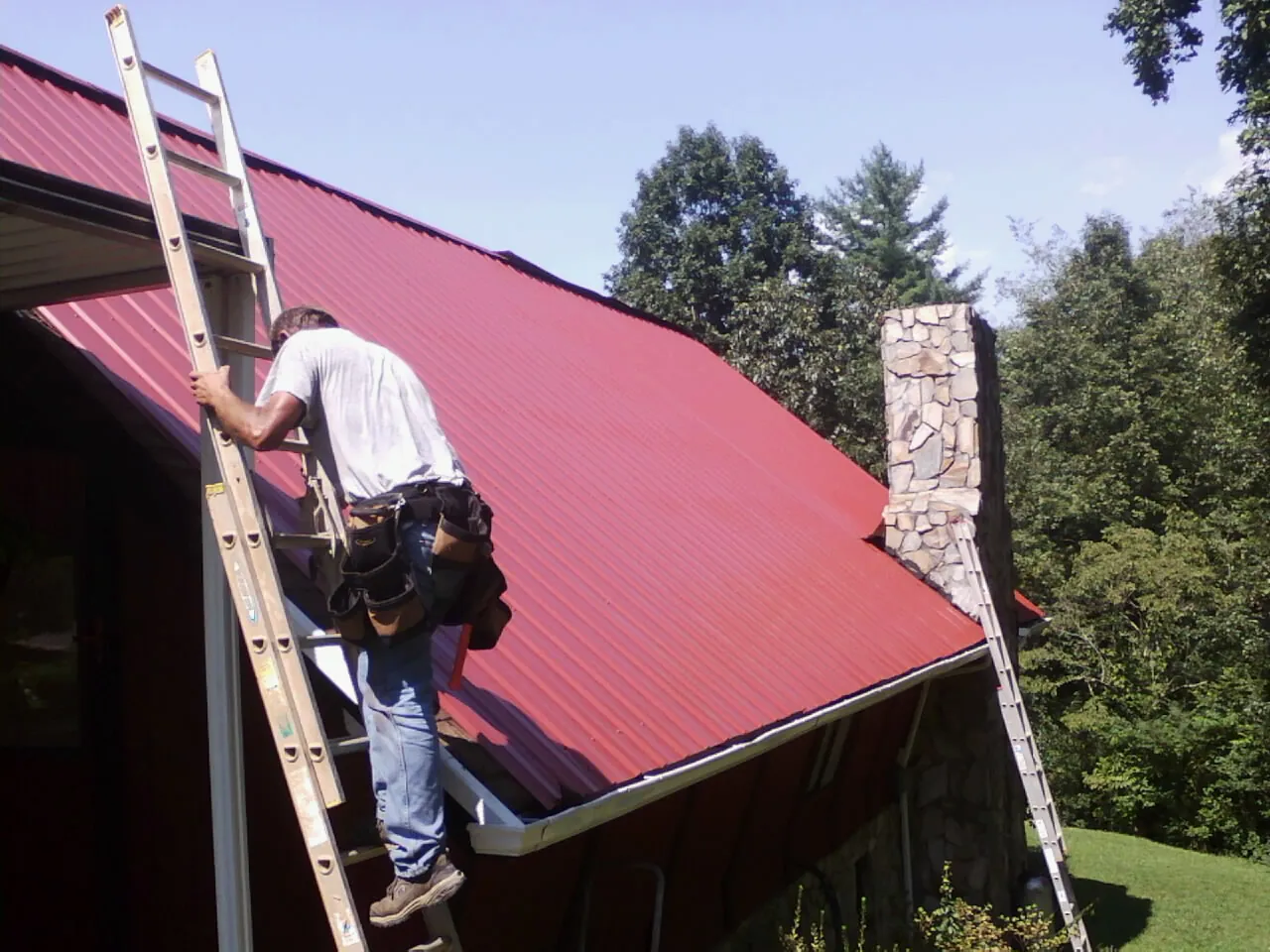 Roofer on a ladder beside a red metal roof and stone chimney — Soffit & Fascia Repair by Roofing Brothers in New Glarus